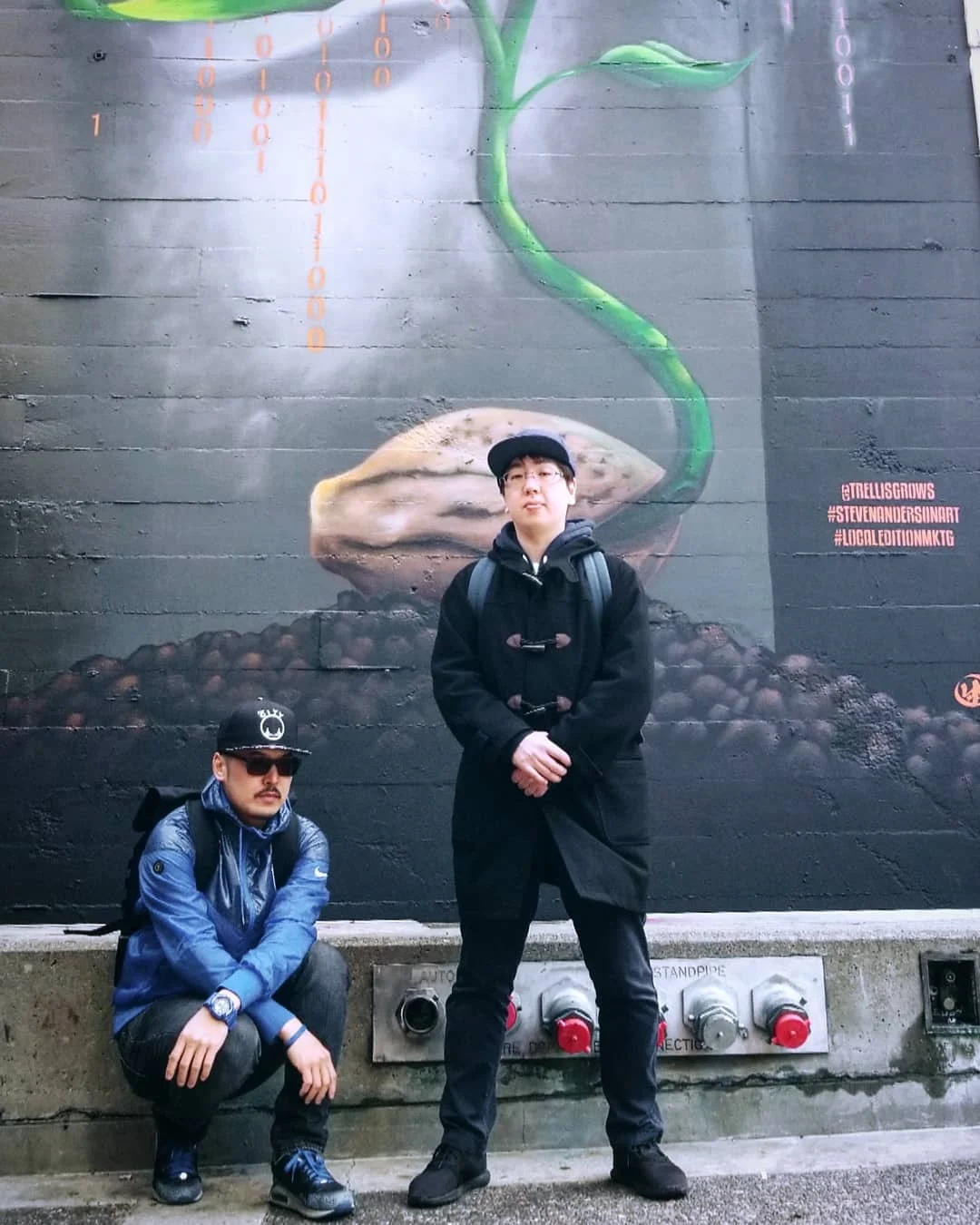 Two people pose below a dark mural of a green sprout growing from a seed, one crouching in a blue jacket and one standing in a black coat.