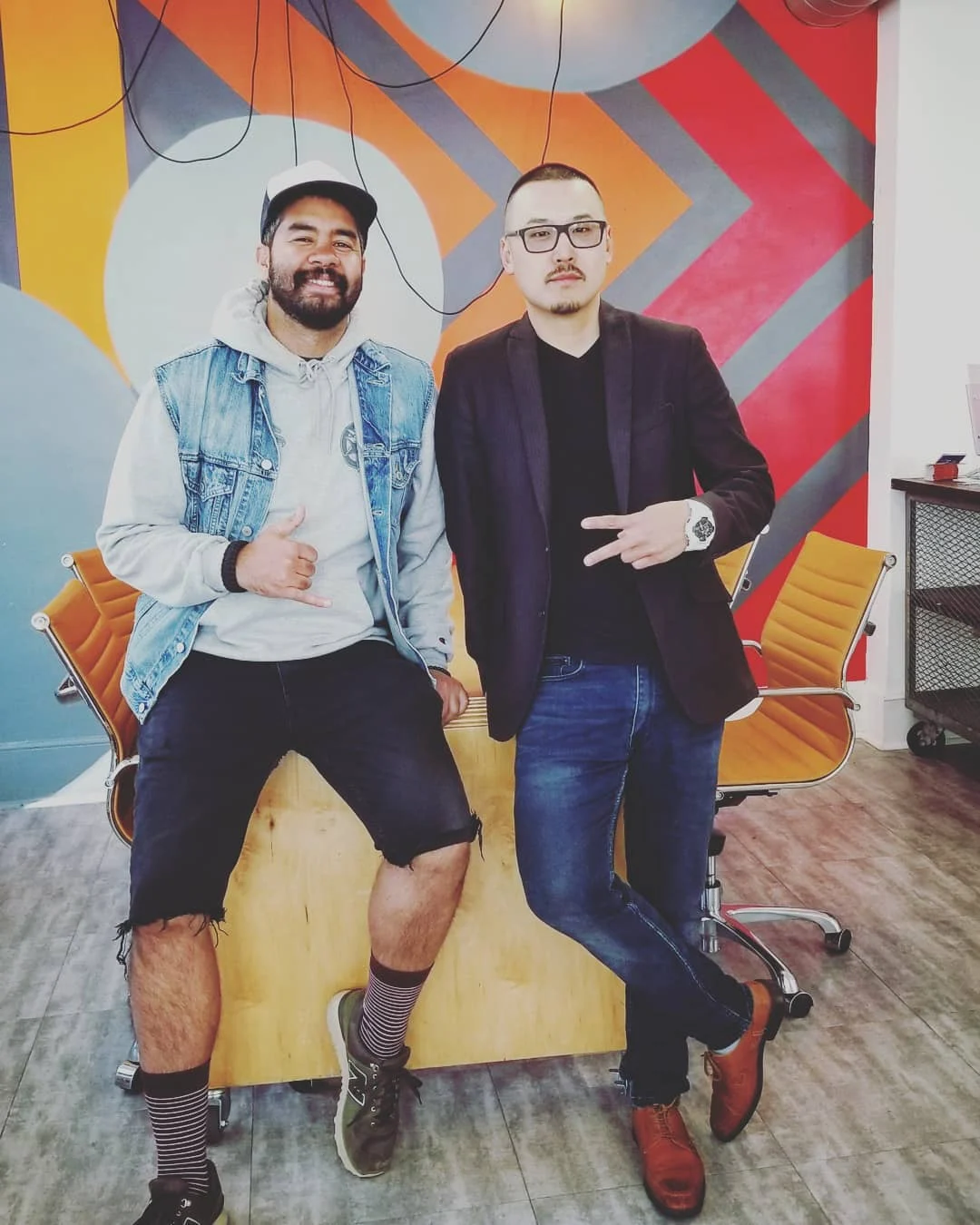 A bearded man in a denim vest and cap and a man in a dark blazer pose in front of a colorful geometric mural.