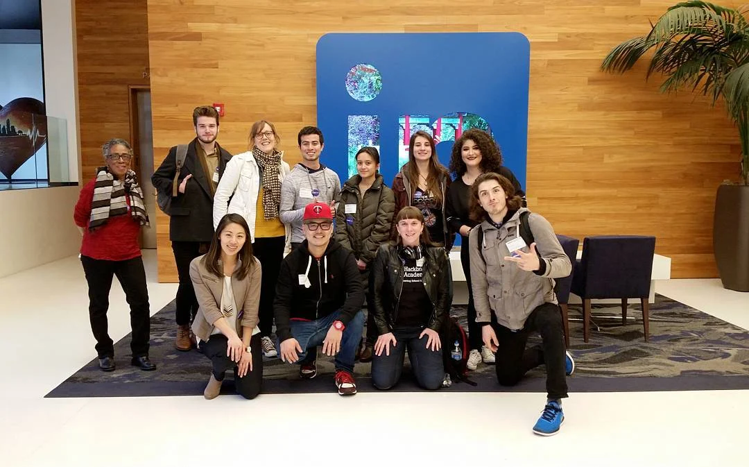Ten people pose in a wood-paneled lobby, some standing and some kneeling, in front of a large blue LinkedIn sign and a potted palm.