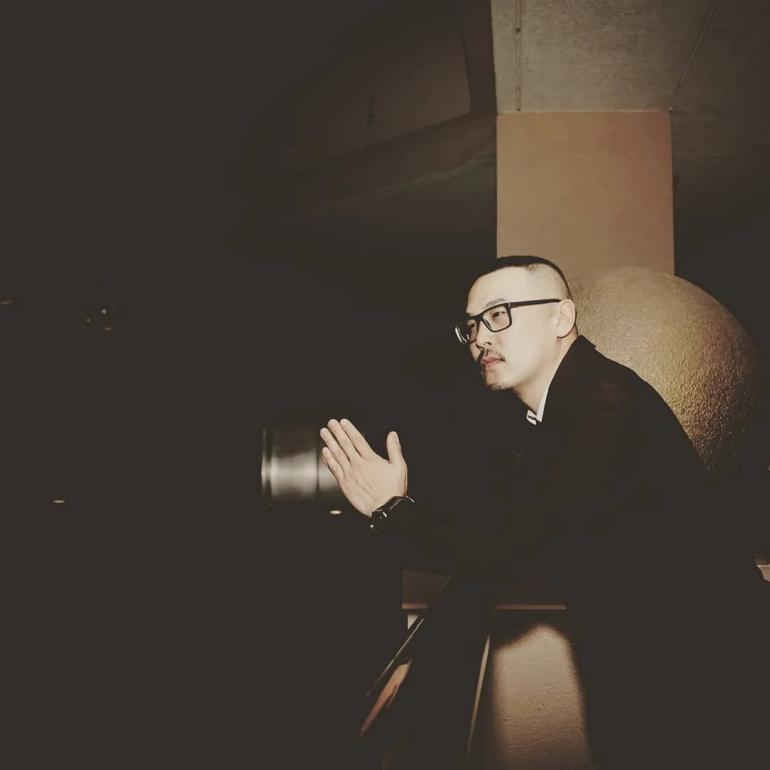 A man with glasses and a short haircut sits in deep shadow, hand raised, lit by a warm glow against a pale pillar.