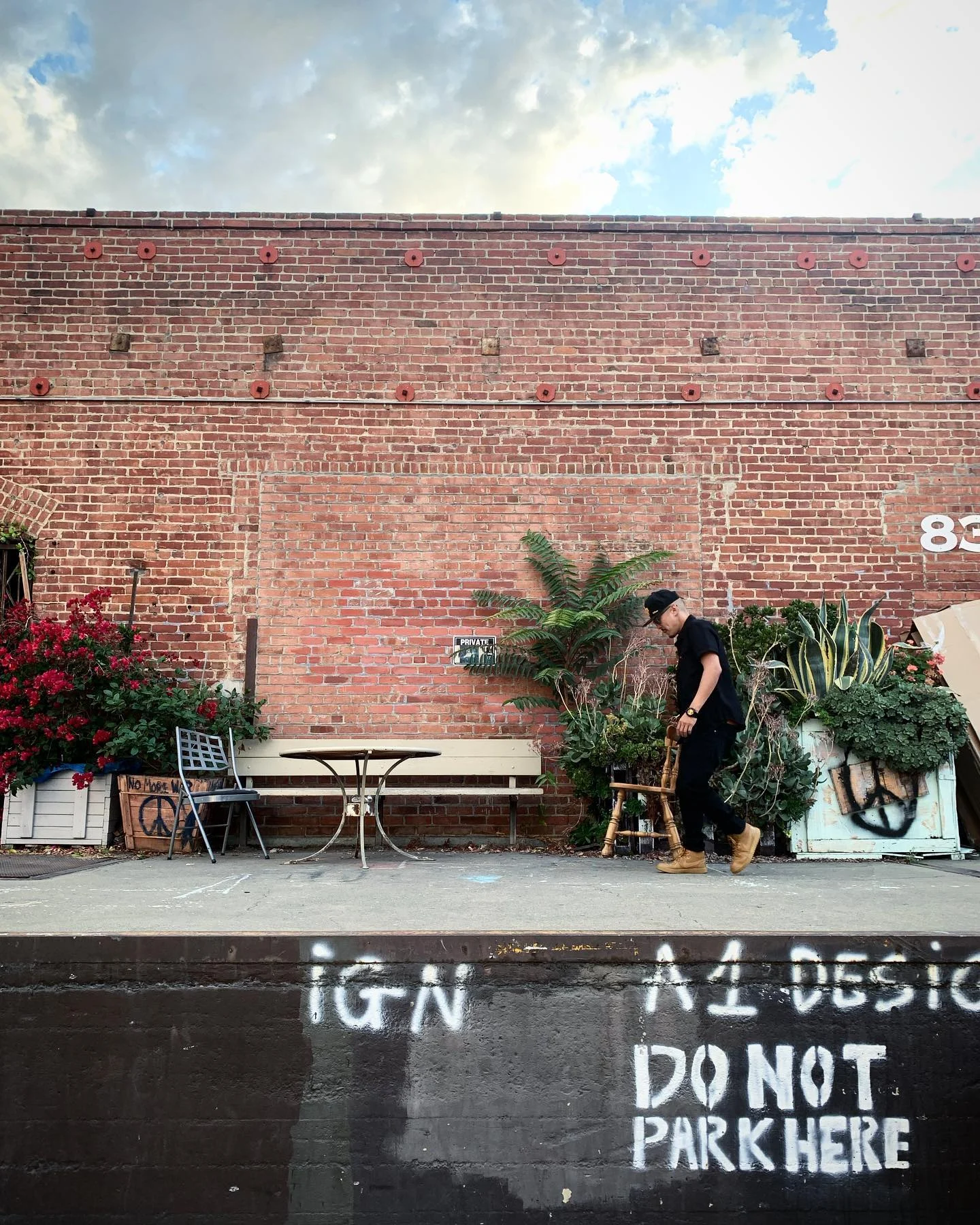 A person in black carries a wooden chair past a red brick wall lined with plants, a bench and flowers, under a cloudy sky.