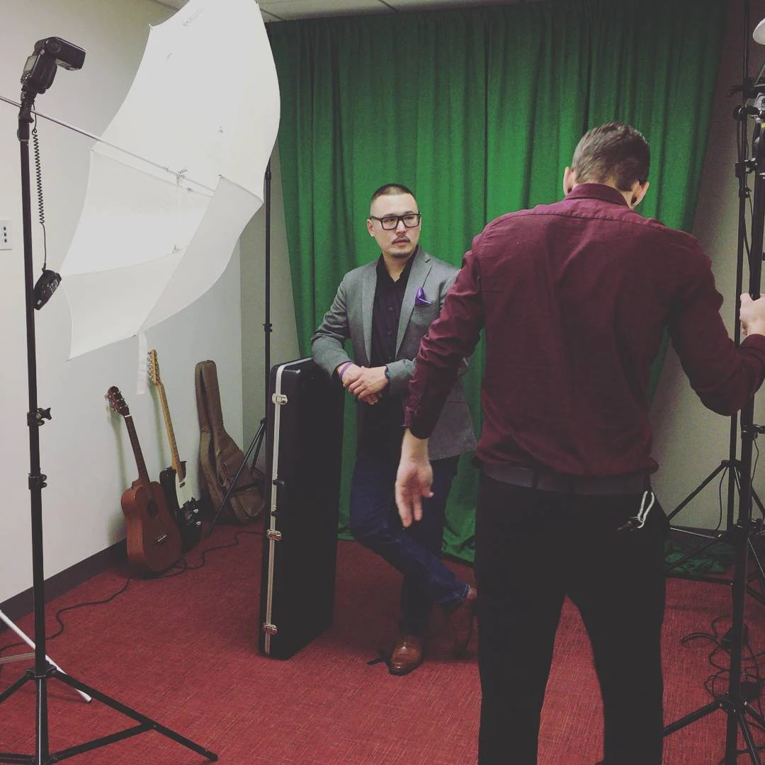 A man in a gray blazer stands before a green curtain beside a guitar case while another man in maroon adjusts studio lights.