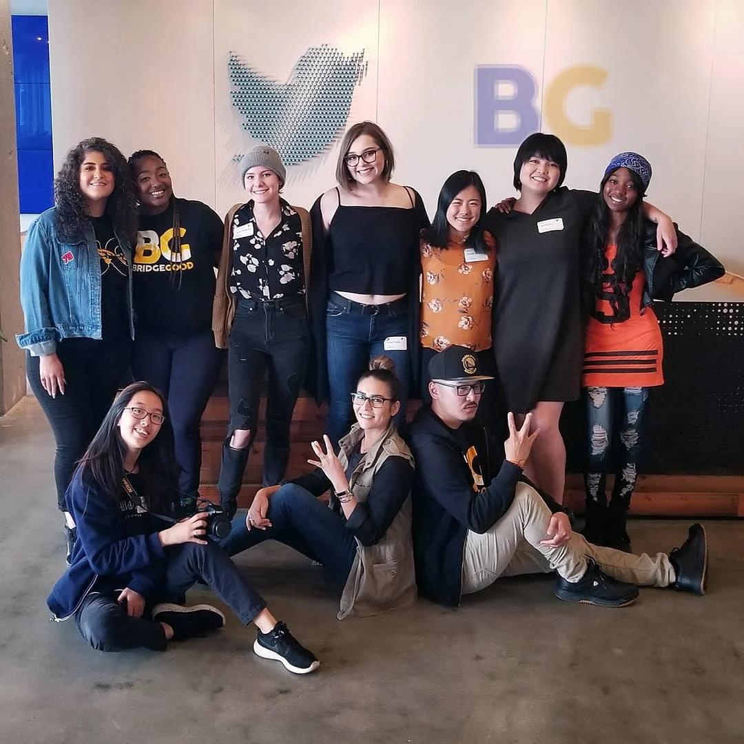Nine people pose against a white wall with the Twitter bird and a BG logo, some seated on the floor making peace signs.