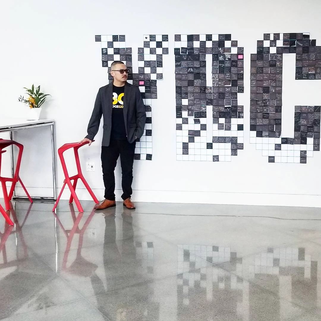 A man in sunglasses and a gray blazer stands by red stools in front of a wall of black and white pixel squares.