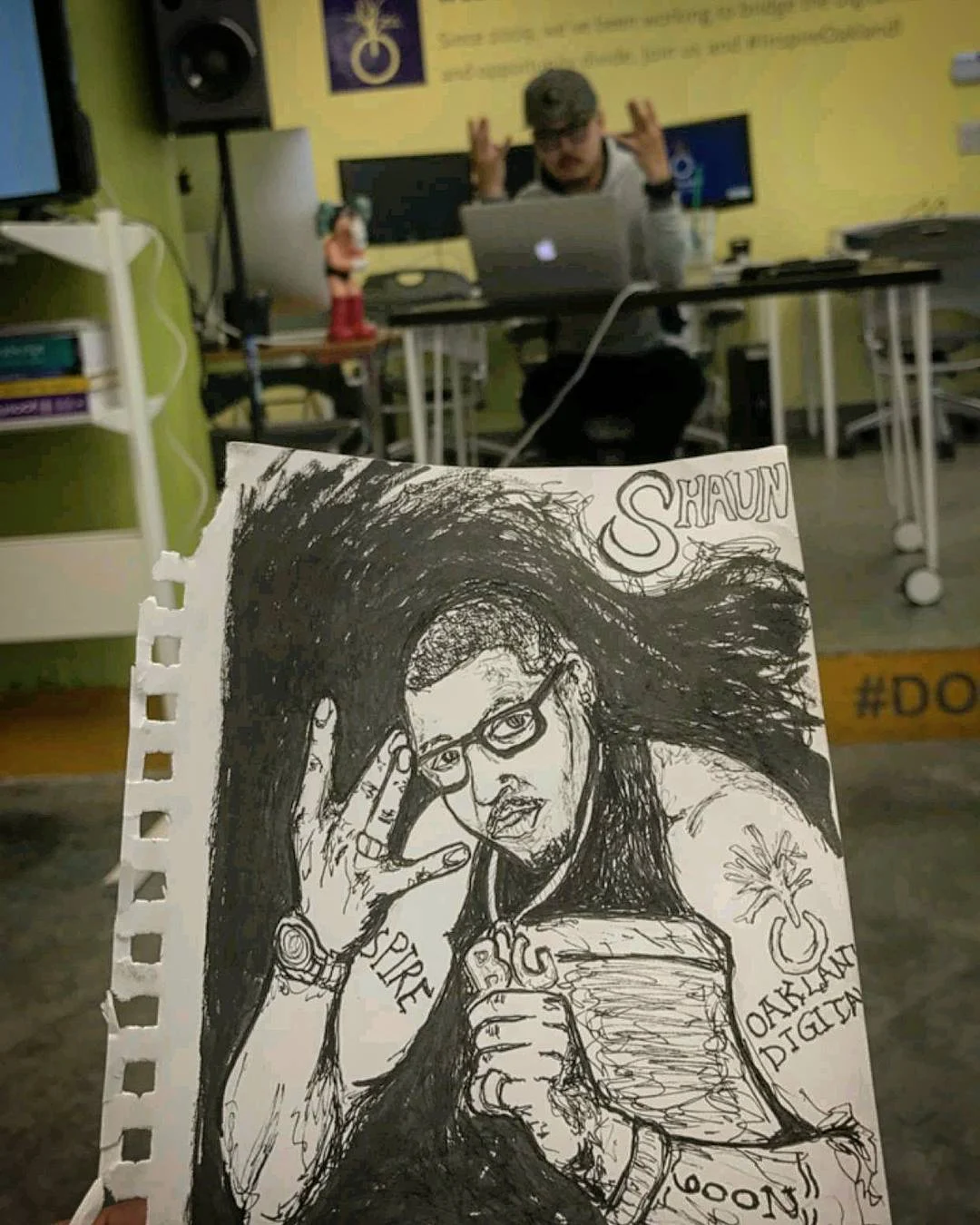 A hand holds up a black pen portrait of a man with glasses, with a man at a laptop in a yellow classroom behind it.