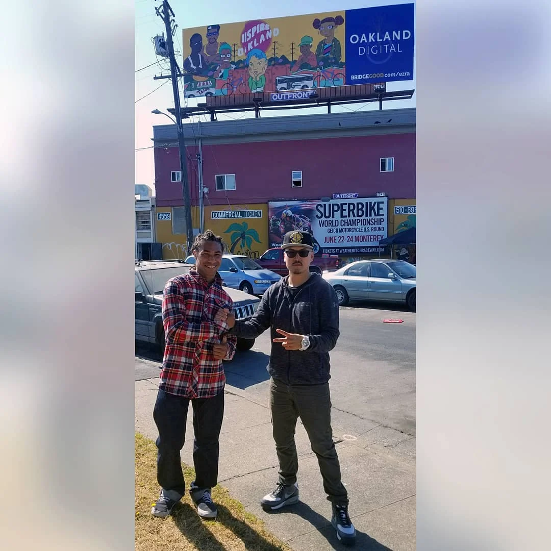 Two men shake hands on a sidewalk under a colorful Oakland Digital billboard, with parked cars and a red building behind them.