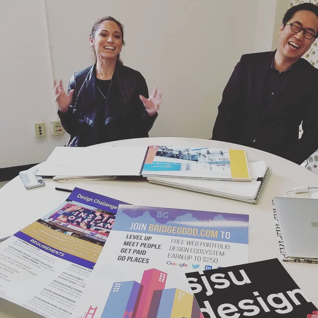 A woman and a man laugh at a round table covered with open portfolios, design flyers and a black sjsu design sign.