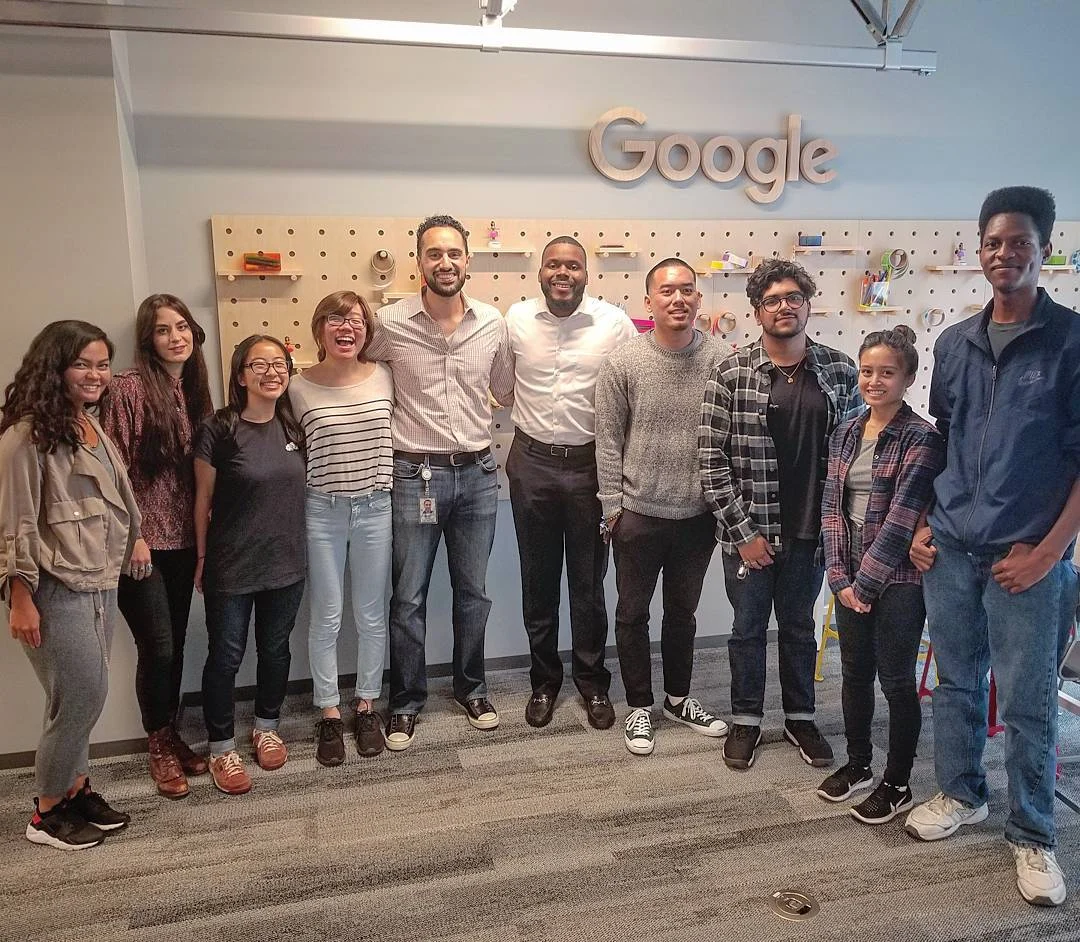 Eleven smiling people stand in a row under a Google sign in front of a pegboard wall in a gray office.