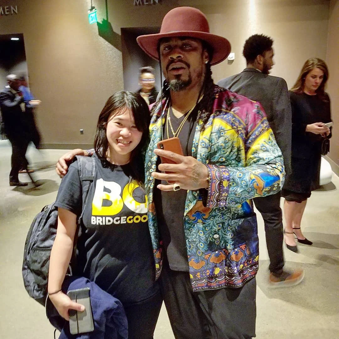 A smiling woman in a BRIDGEGOOD shirt stands beside a man in a maroon hat and colorful patterned jacket in a hallway.