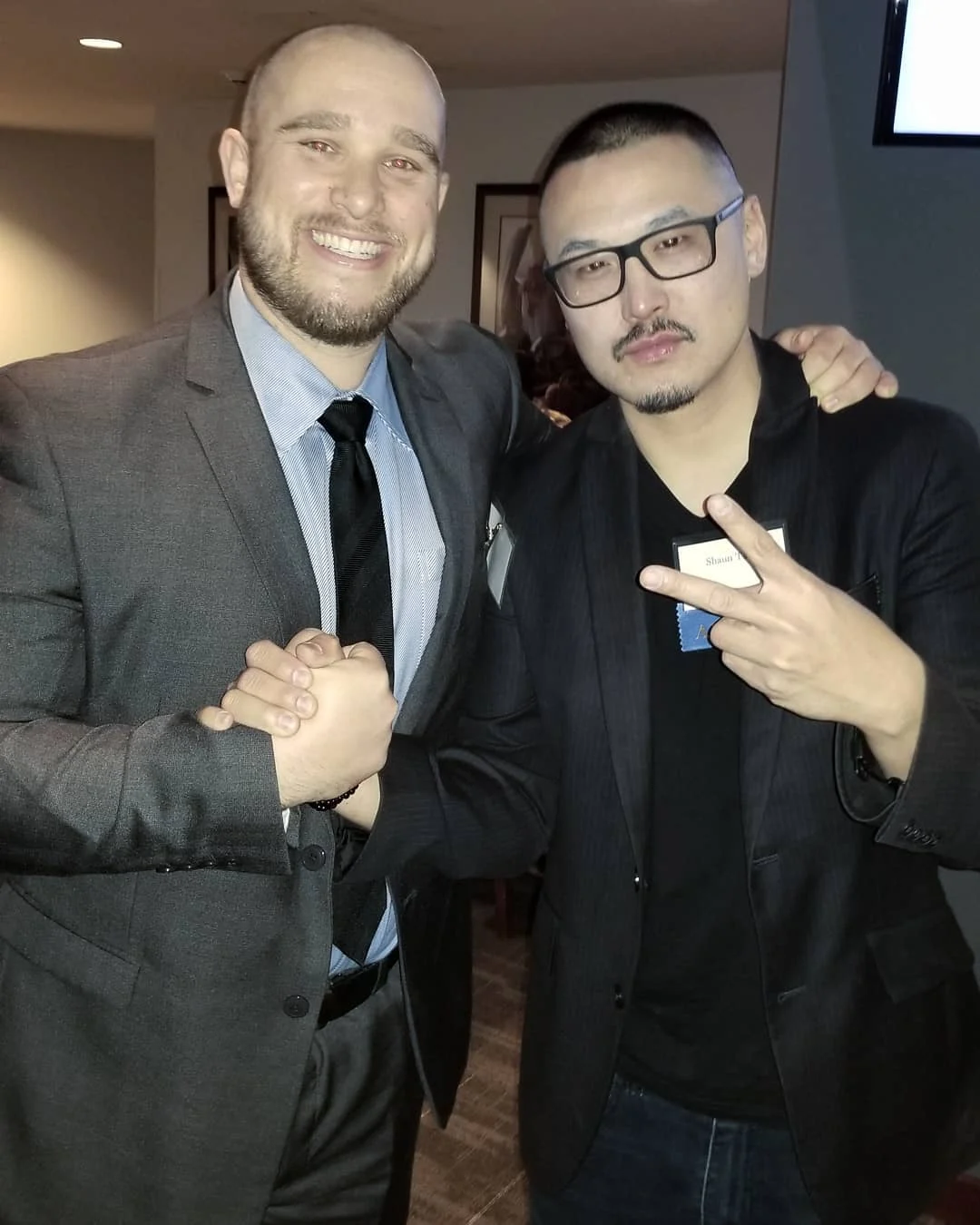 Two men in dark jackets clasp hands and smile in an indoor venue, one making a peace sign with a name tag on his lapel.