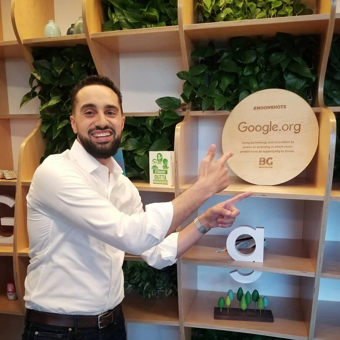 A smiling bearded man in a white shirt points at a round wooden Google.org sign on shelves with green plants.