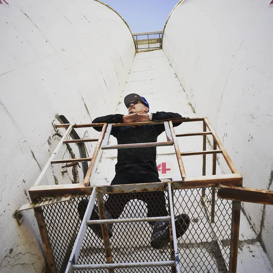 A man in sunglasses and a cap leans on a rusty platform at the top of a ladder between two tall curved white walls.