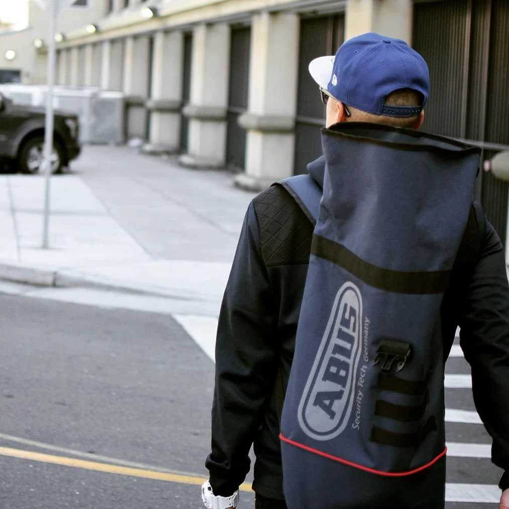 A person in a blue cap and black jacket, seen from behind, crosses a street with a long navy ABUS bag on their back.