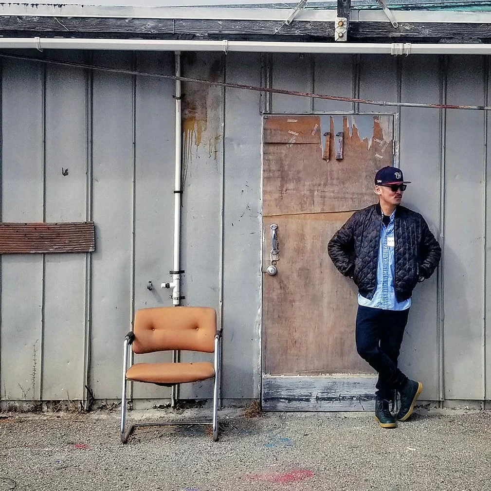 A man in a cap and quilted jacket leans on a weathered door beside an orange chair on gravel outside a gray wall.