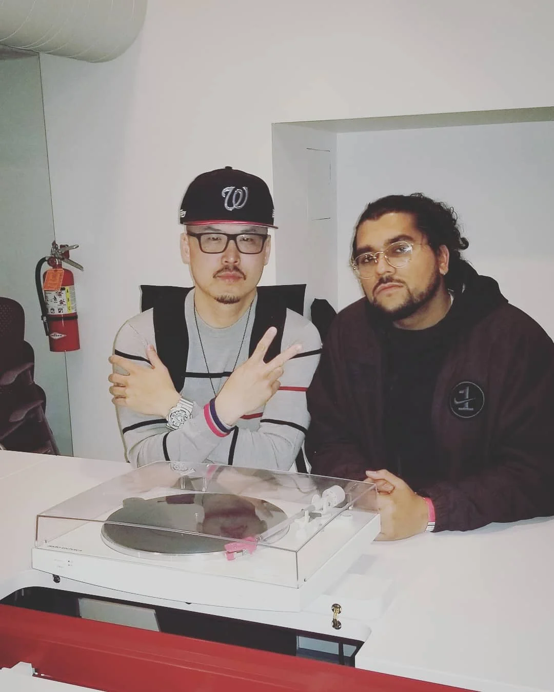 Two men in glasses pose behind a white counter with a turntable, one in a W cap making a peace sign.