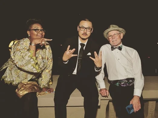 Three people pose on a stone ledge at night, a woman pointing, a man in a tie making hand signs, and a man in a straw hat.