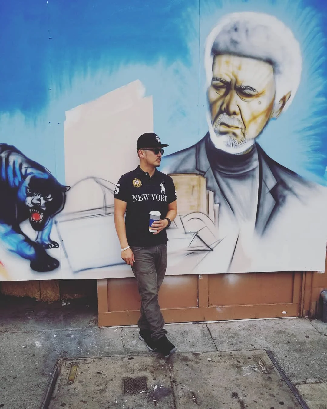 A man in a black cap, sunglasses and polo holds a coffee cup in front of a mural of a panther and an elder.