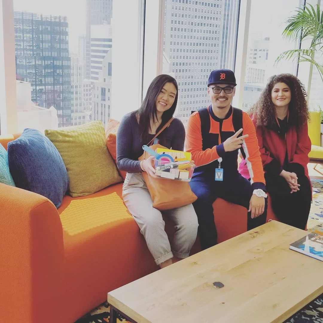 Three smiling people sit on an orange couch with colorful pillows in front of tall windows overlooking city high-rises.