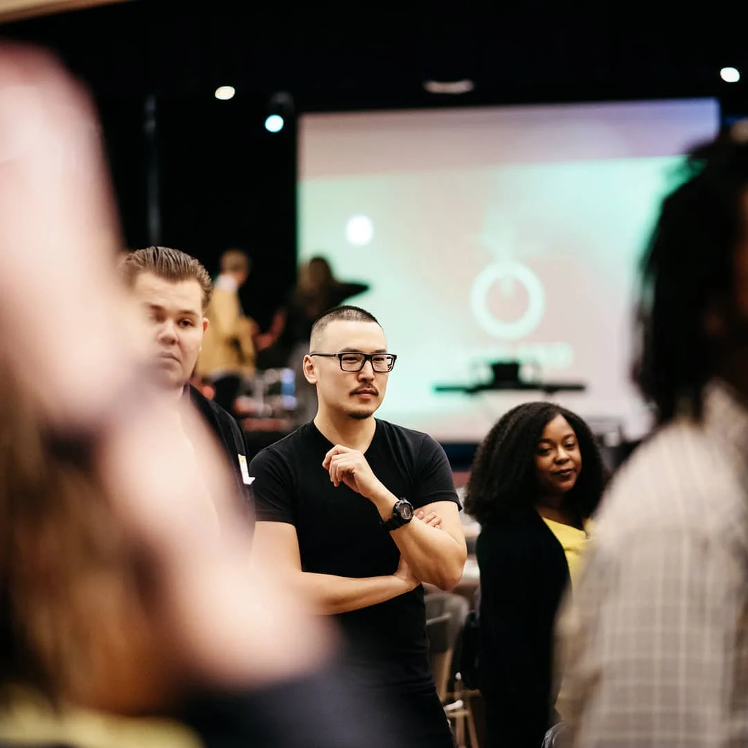 A man with glasses and crossed arms stands among blurred attendees in a dim hall with a glowing projection screen behind.