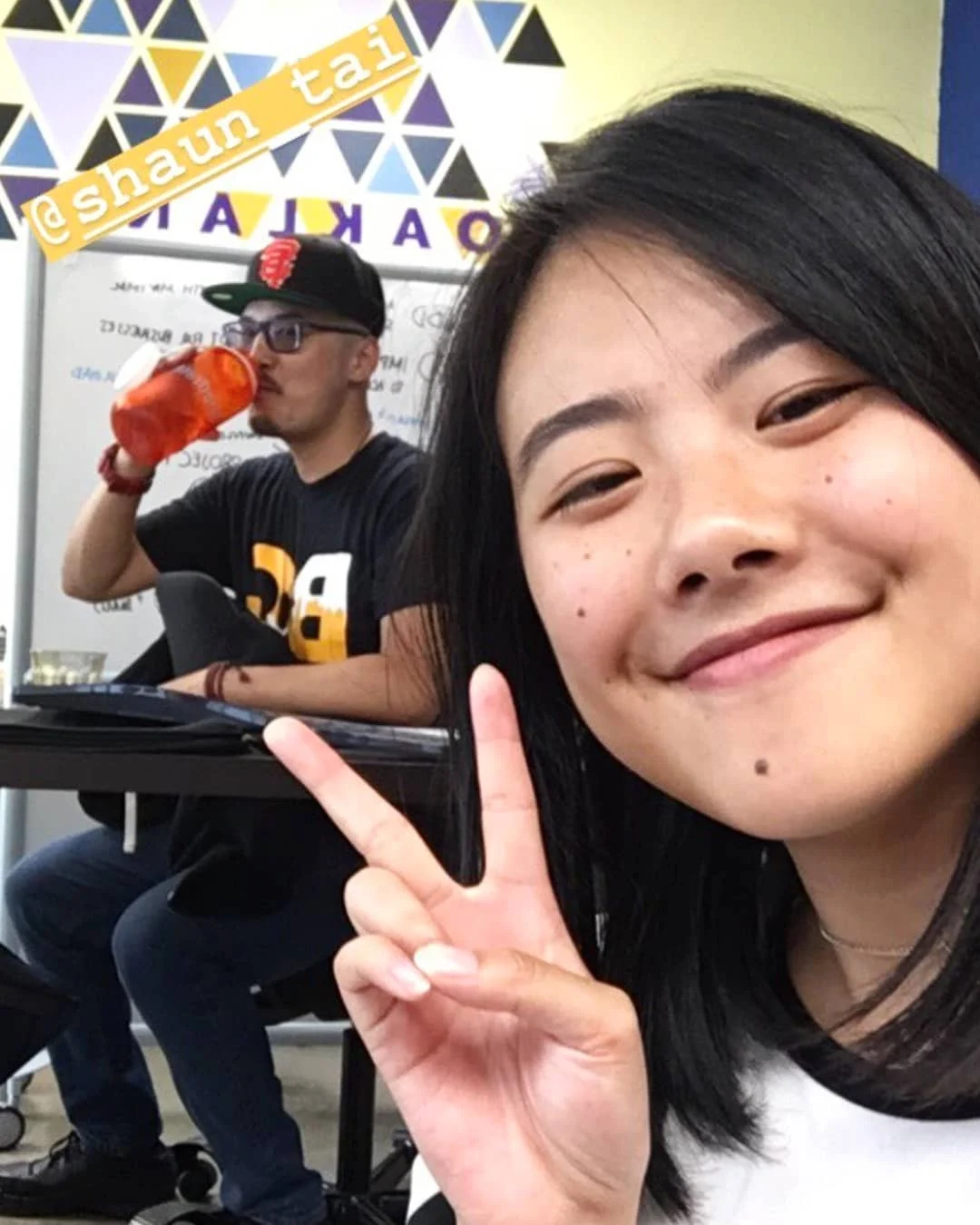 A smiling woman makes a peace sign in a selfie while a man in a cap and black shirt drinks behind her.
