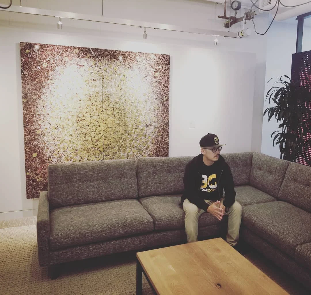 A man in a black BG hoodie sits on a gray sectional couch beneath a large gold and brown canvas.