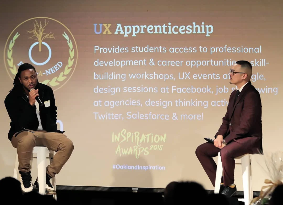 Two men sit on white stools onstage, one holding a microphone, before a projected slide titled UX Apprenticeship.