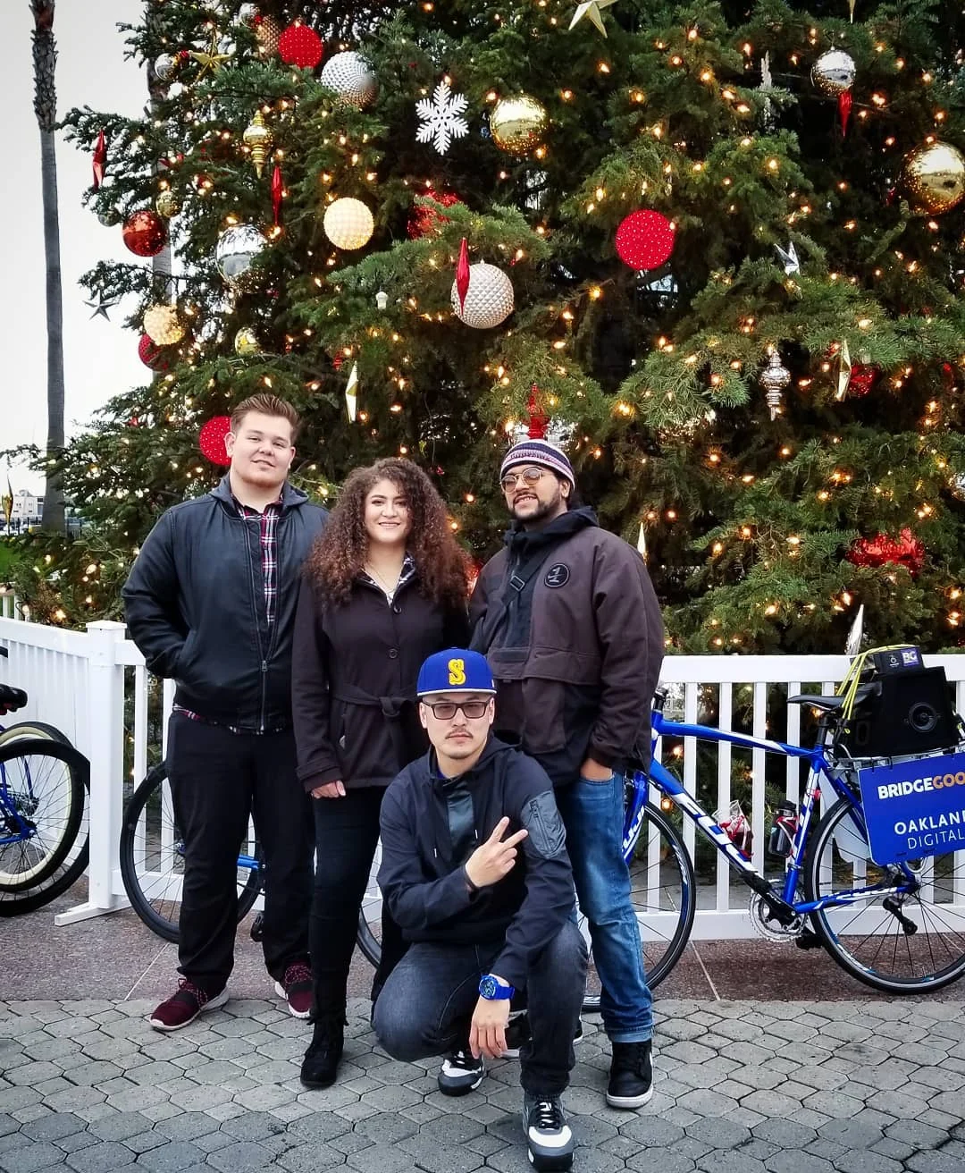 Four people pose in front of a tall Christmas tree with ornaments, with blue bikes behind a white fence.