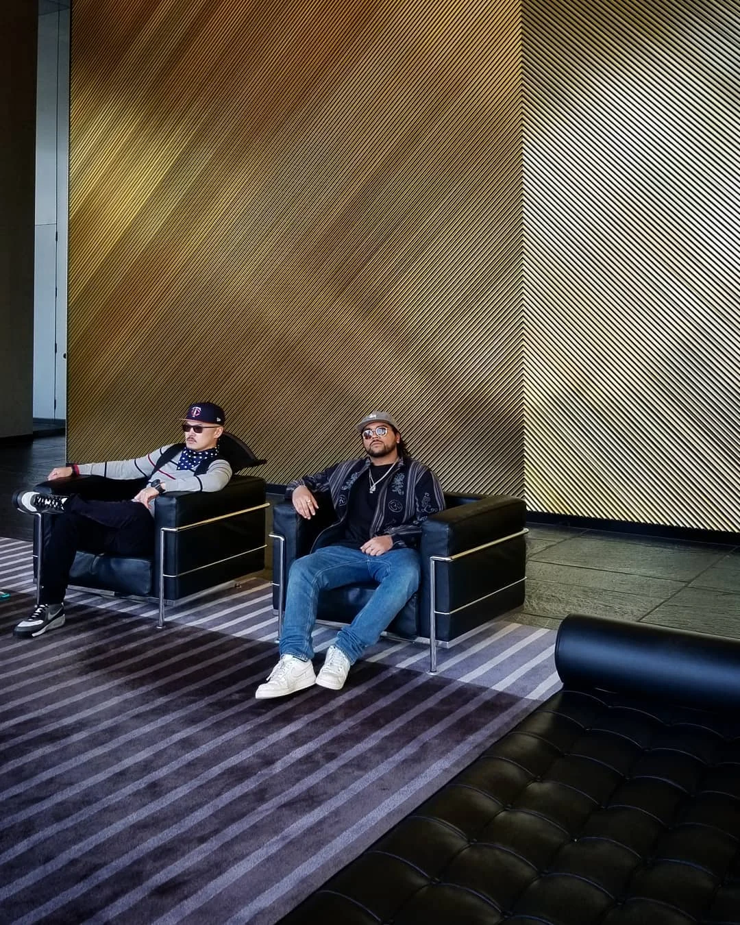Two men in caps sit in black leather armchairs in front of a gold striped wall and a gray striped rug.
