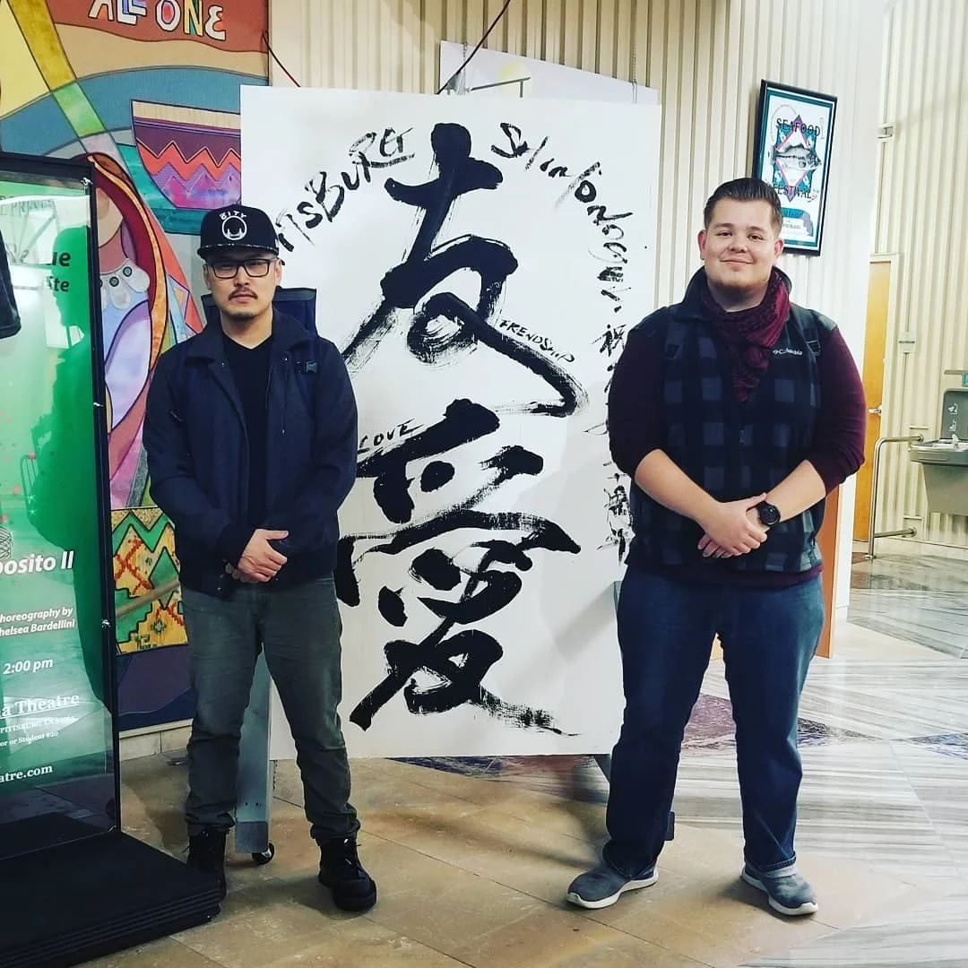Two men stand beside a large white banner with black brush calligraphy in a lobby with a colorful mural.