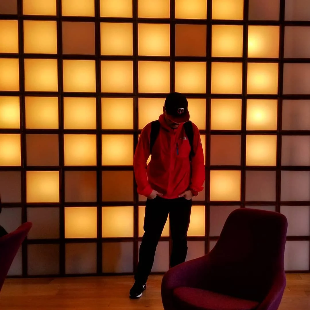 A person in a red jacket and black cap stands before a wall of glowing orange squares beside a plum chair.