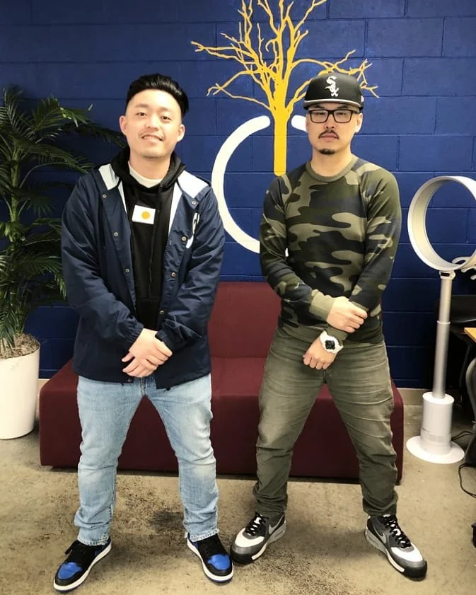 Two men pose in front of a blue brick wall with a yellow tree, one in a navy jacket and one in camo.