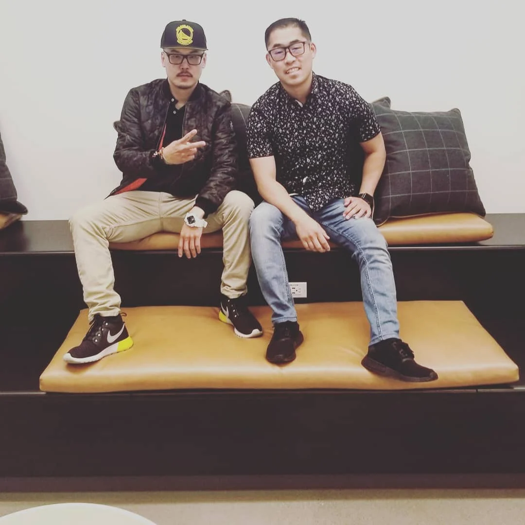 Two men sit on tan leather cushions on a tiered bench, one in a cap making a peace sign.