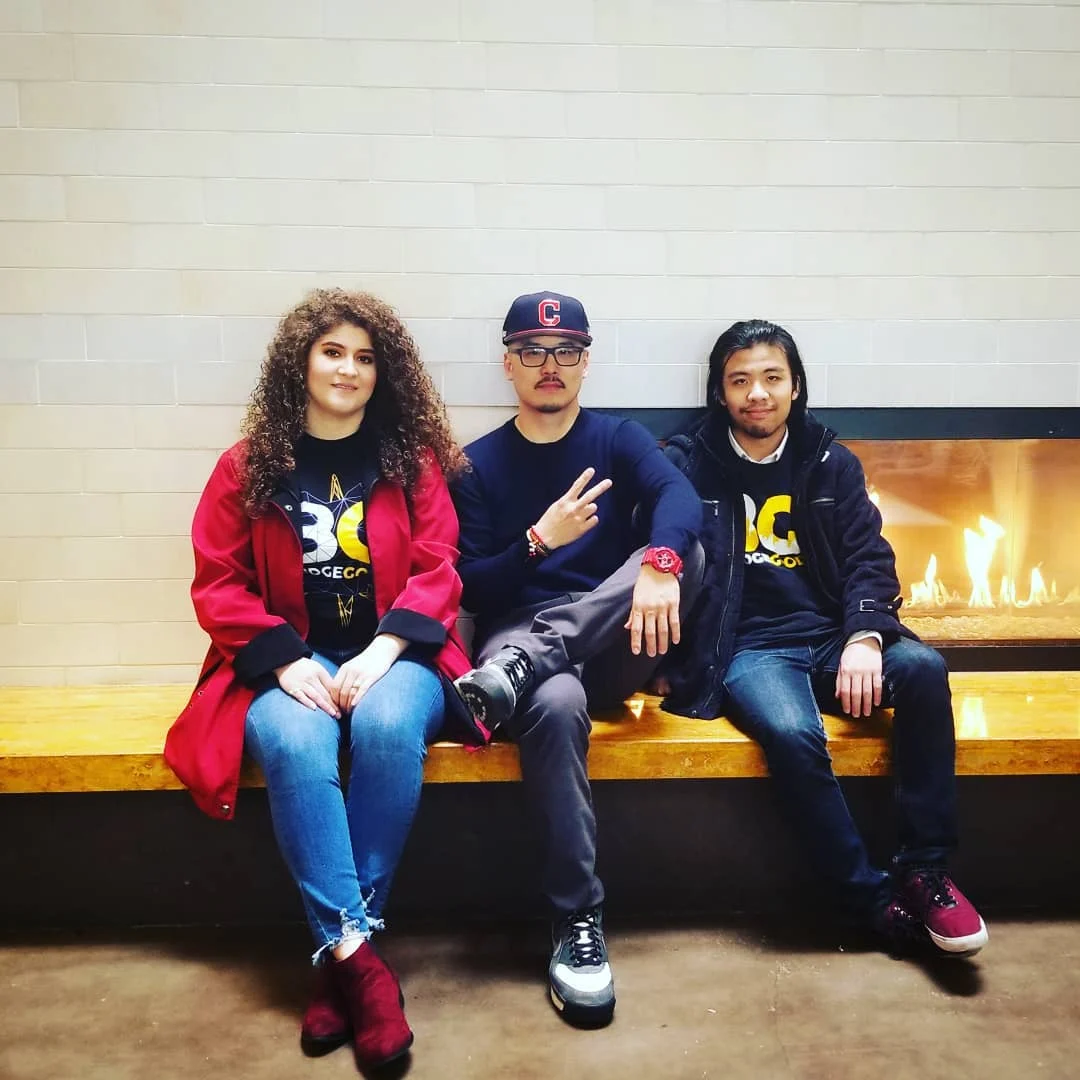 Three people sit on a wooden bench beside a lit fireplace, two in BG shirts and one making a peace sign.