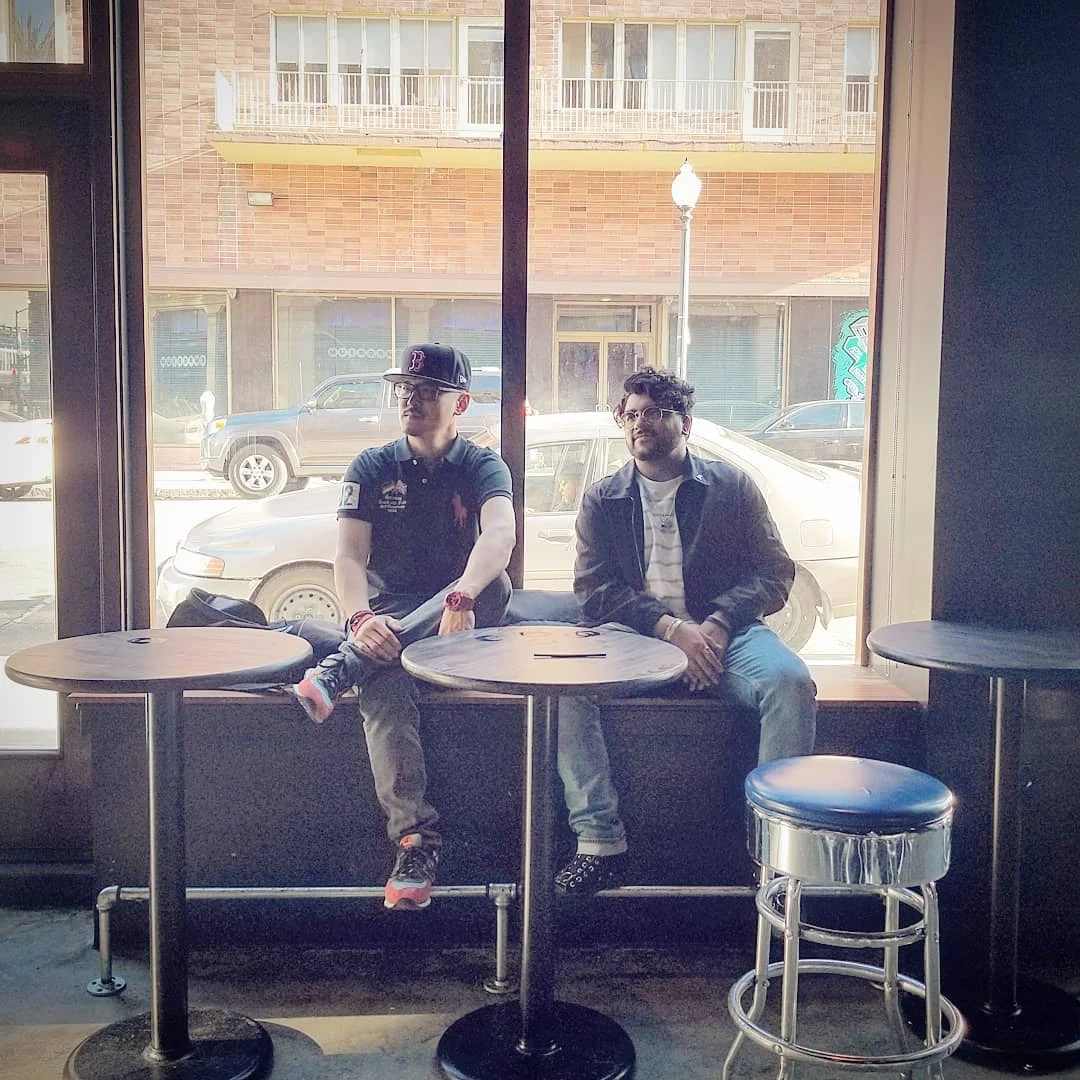 Two men sit on a wide window ledge between round tables, with a blue stool and a street outside.