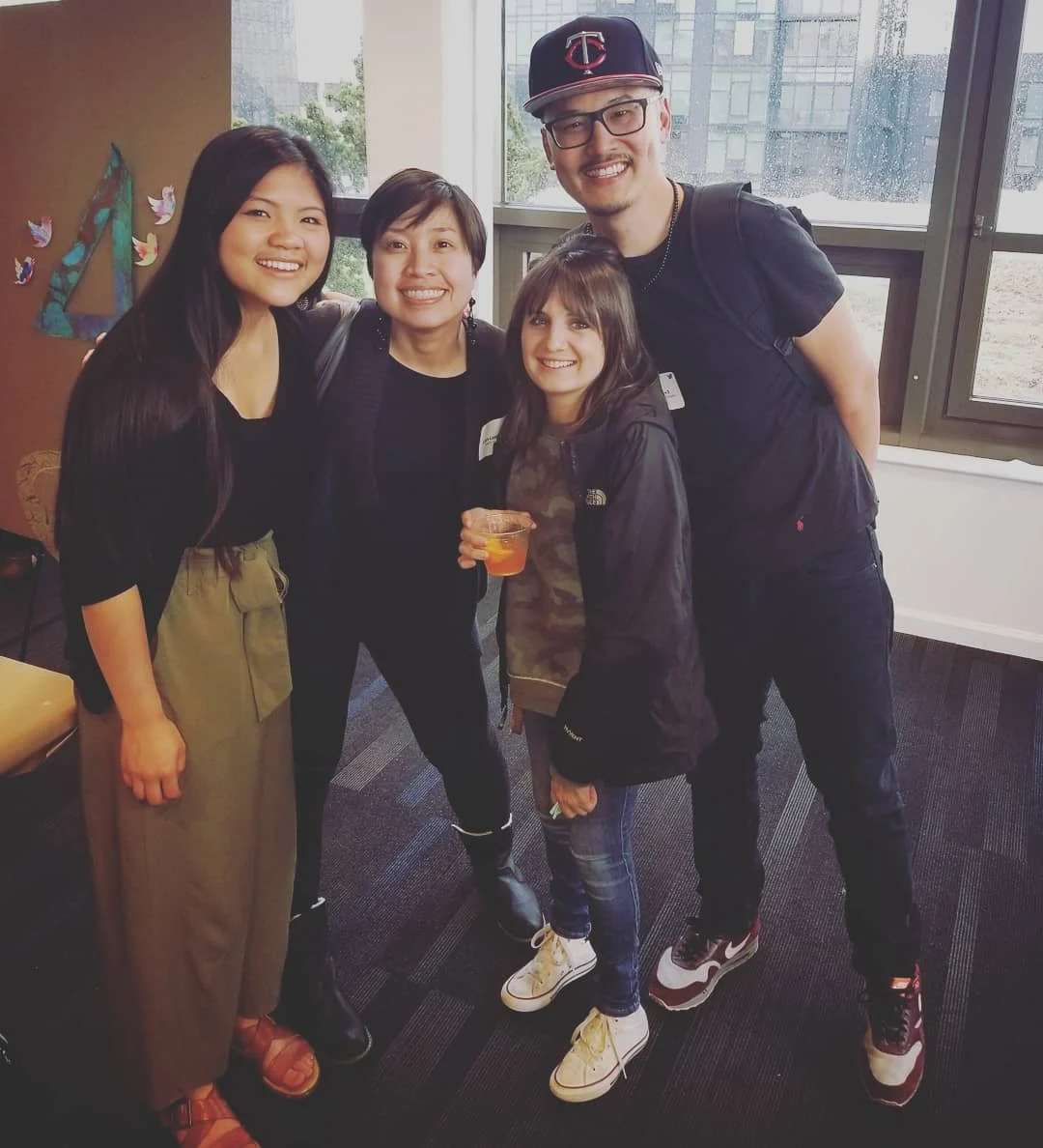 Four smiling people crowd together beside large windows, one holding an orange drink in a clear cup.