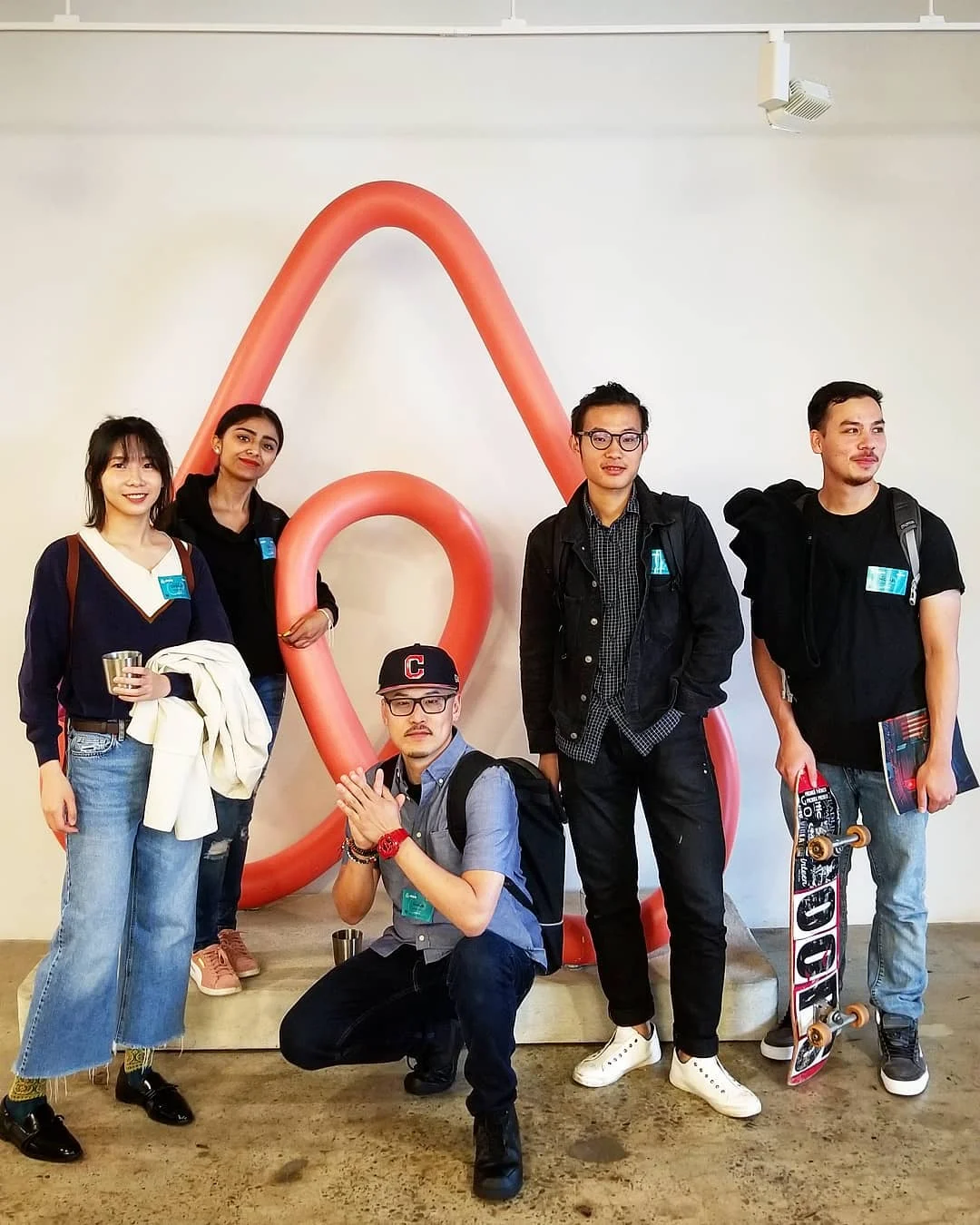 Five people with name stickers pose before a large coral Airbnb logo sculpture, one holding a skateboard.