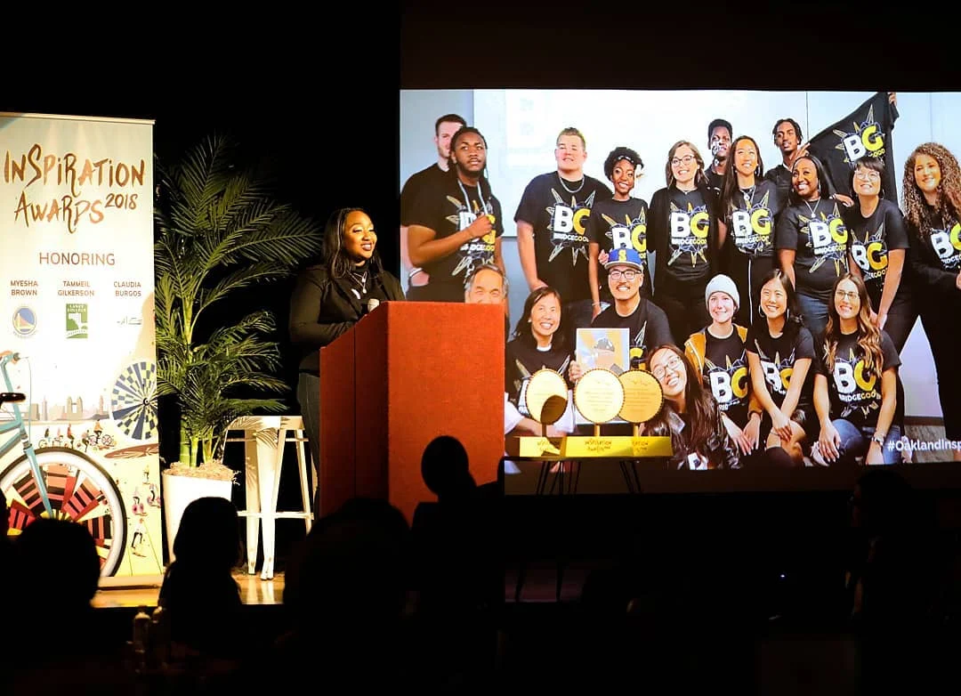 A woman speaks at a podium on a dark stage beside a screen showing a large group in BG shirts.