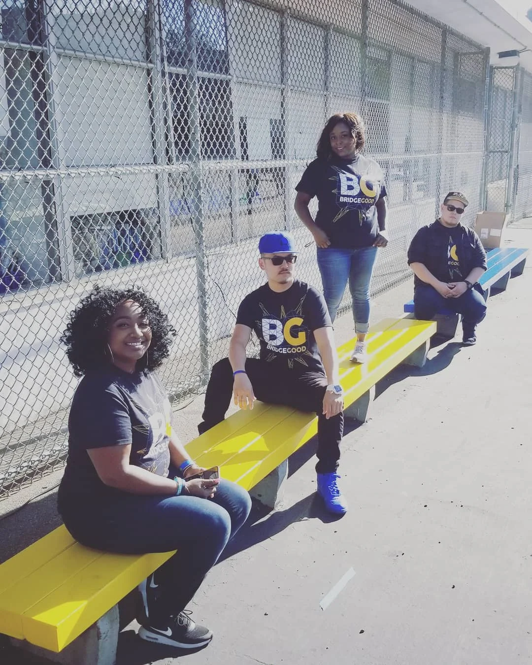 Four people in black BG shirts sit and stand on yellow benches beside a chain-link fence in sunlight.