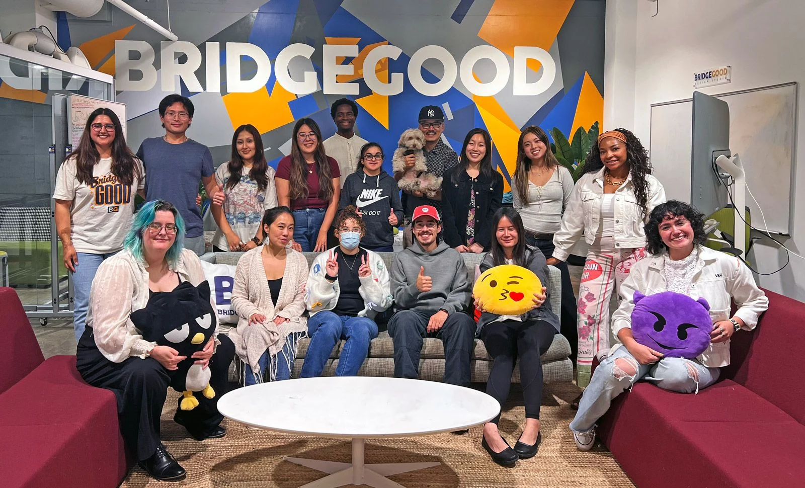 A group poses in front of a BRIDGEGOOD mural with white letters, orange and blue shapes, and red chairs.