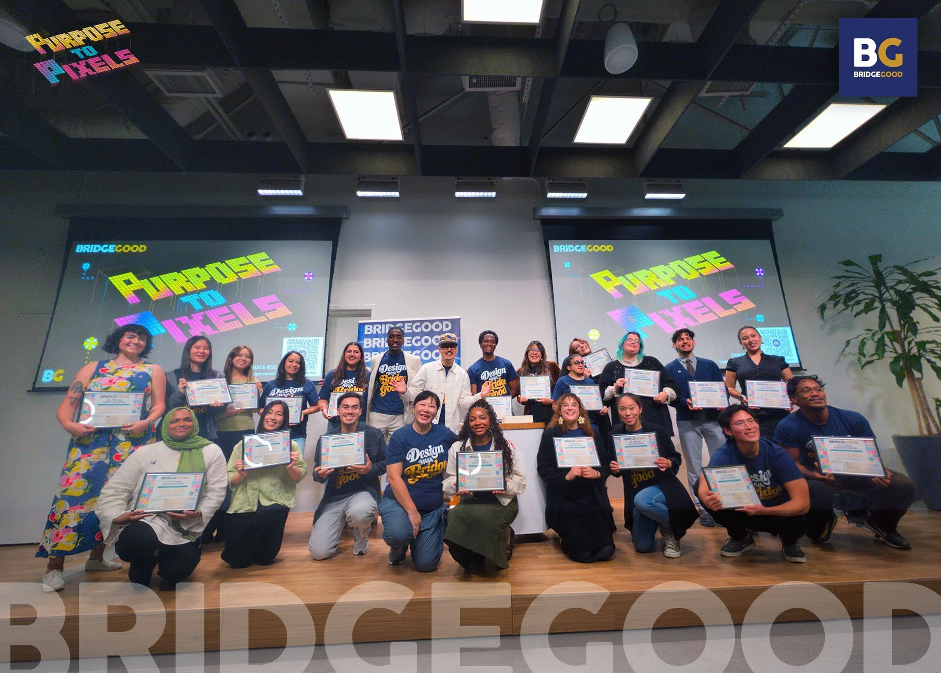 A group in blue shirts kneels and stands on a wooden stage holding certificates before Purpose to Pixels screens and a BridgeGood banner.