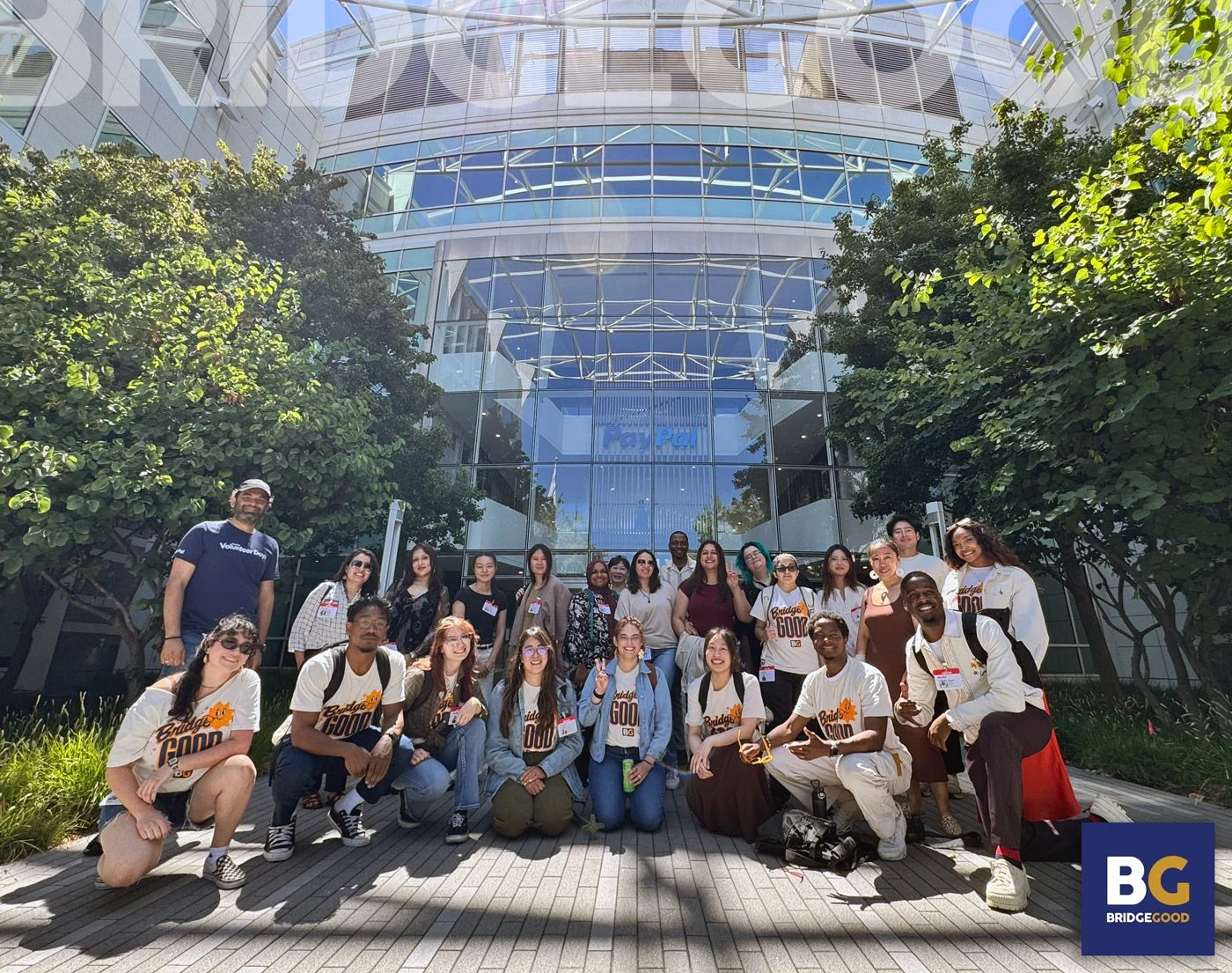 A large group of people in matching white BridgeGood shirts poses outdoors in front of a glass building surrounded by green trees.