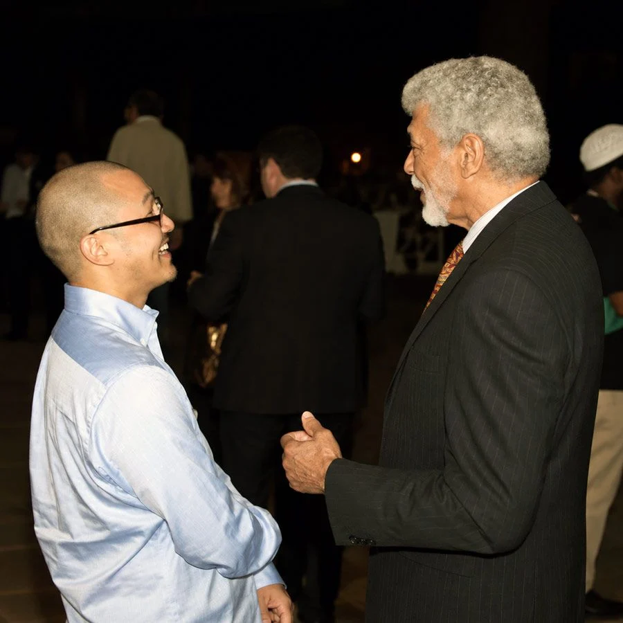Two men stand facing each other at a dim indoor event, one in a light blue shirt and glasses laughing, the other in a dark pinstripe suit gesturing with his hand.