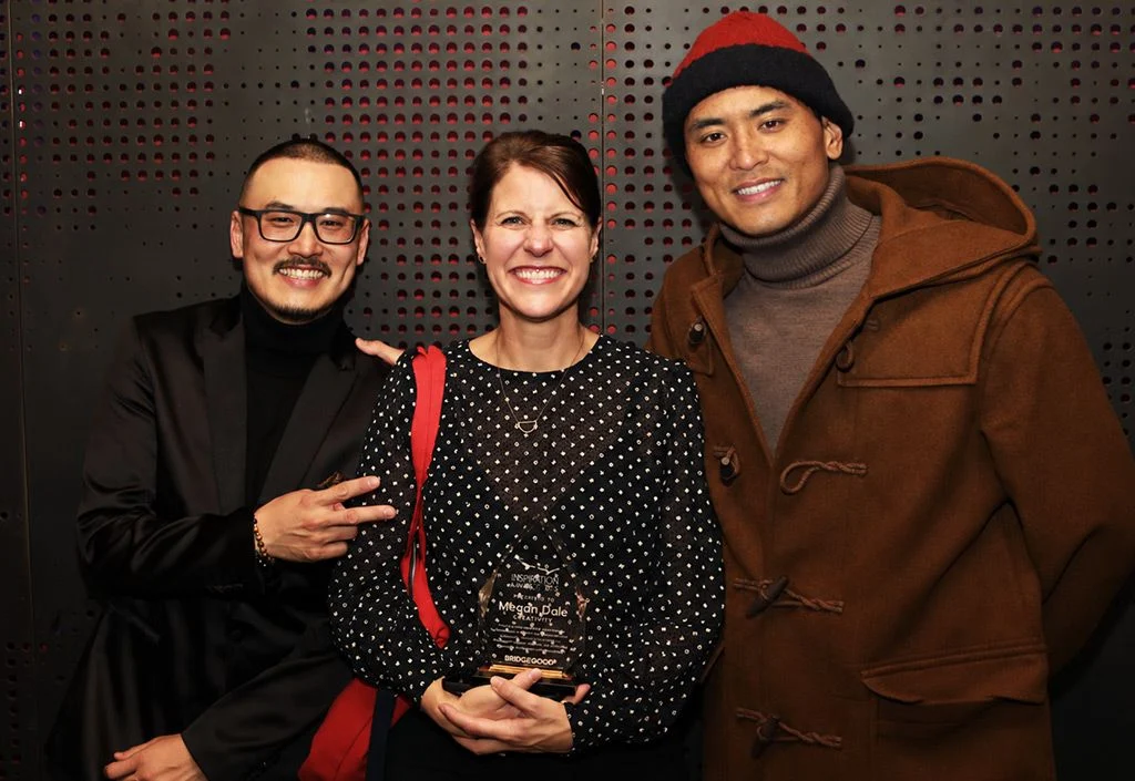Three people smile together in front of a dark perforated metal wall with red accents, one holding a glass award.