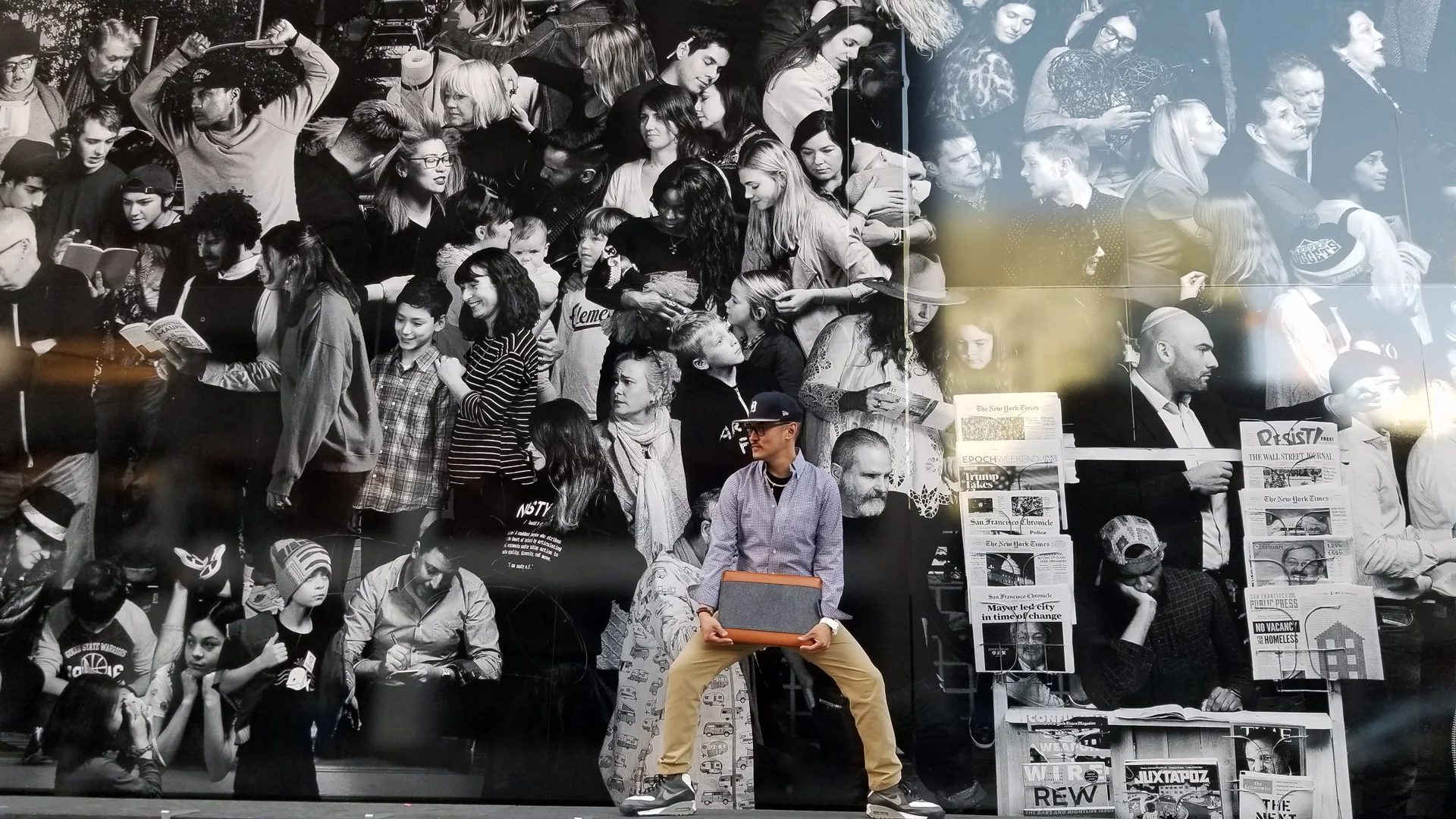A man in a lavender shirt crouches holding a brown box in front of a black and white crowd mural.