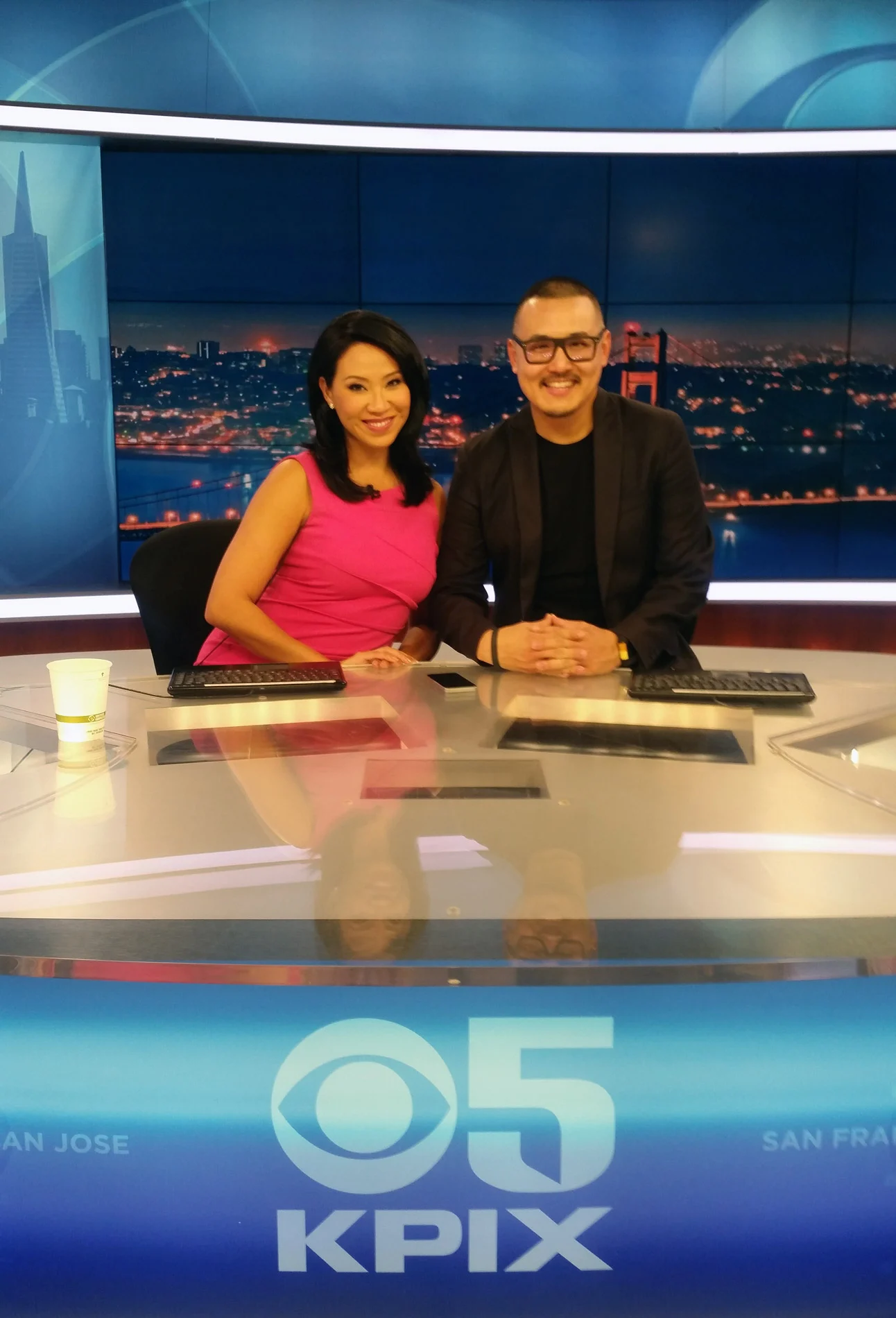 Two smiling people sit behind a glossy news anchor desk in a television studio, with a night city skyline glowing blue behind them.