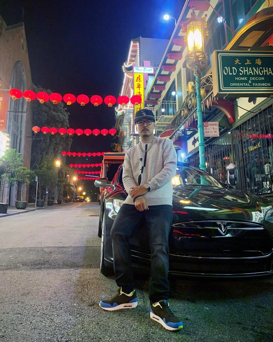A person in a gray sweatshirt and black cap sits on a dark car at night, lit by red lanterns and a street lamp in a city street.