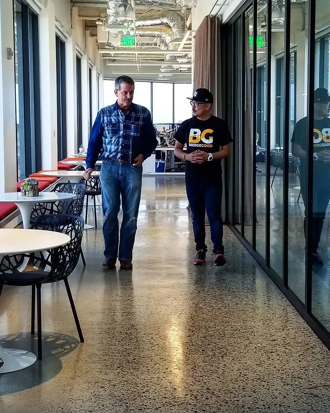 Two men walk side by side down a bright office corridor with polished concrete floors, red-cushioned banquettes, white tables, and glass walls, one in a plaid shirt and jeans, the other in a black BRIDGEGOOD t-shirt and cap.