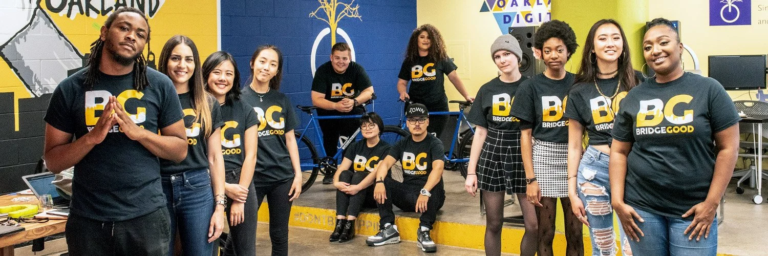A group of people in matching black BridgeGood t-shirts poses in a bright studio with yellow and blue walls, a bicycle behind them.