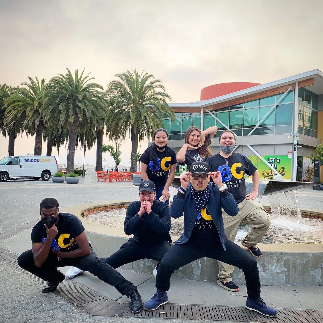A group of six people in matching black shirts pose in a crouch beside a fountain, with palm trees and a glass building behind them.