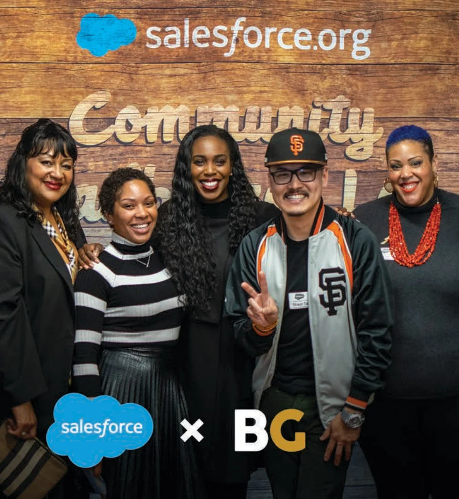 Five people pose smiling before a wooden wall, and a man in a gray jacket flashes a peace sign.